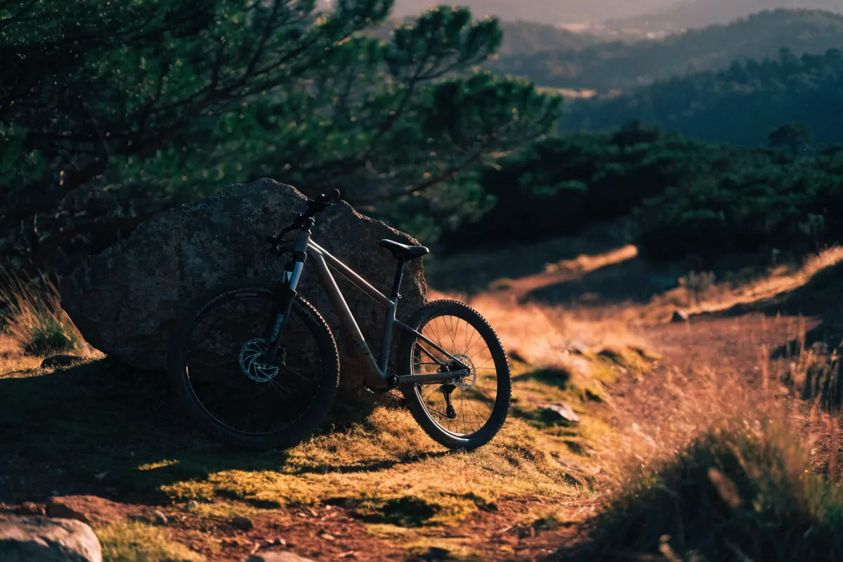 Mountain bike aro 29 apoiada numa pedra em trilha de terra, com montanhas ao fundo no fim da tarde