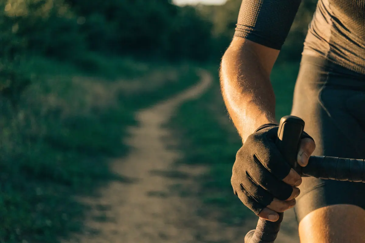 Ciclista de bermuda e luva de dedo curto com a mão no guidão, trilha de terra à frente